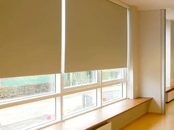 Stores enrouleurs beiges baissés devant les baies d’une salle claire, qui diffusent la lumière