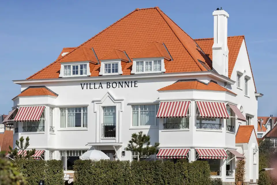 Façade blanche d’un hôtel dont les fenêtres sont abritées par des stores à projection à toile rayée rouge et blanc