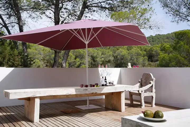 Parasol à toile bordeaux au-dessus d’une longue table en bois, sur une terrasse blanche