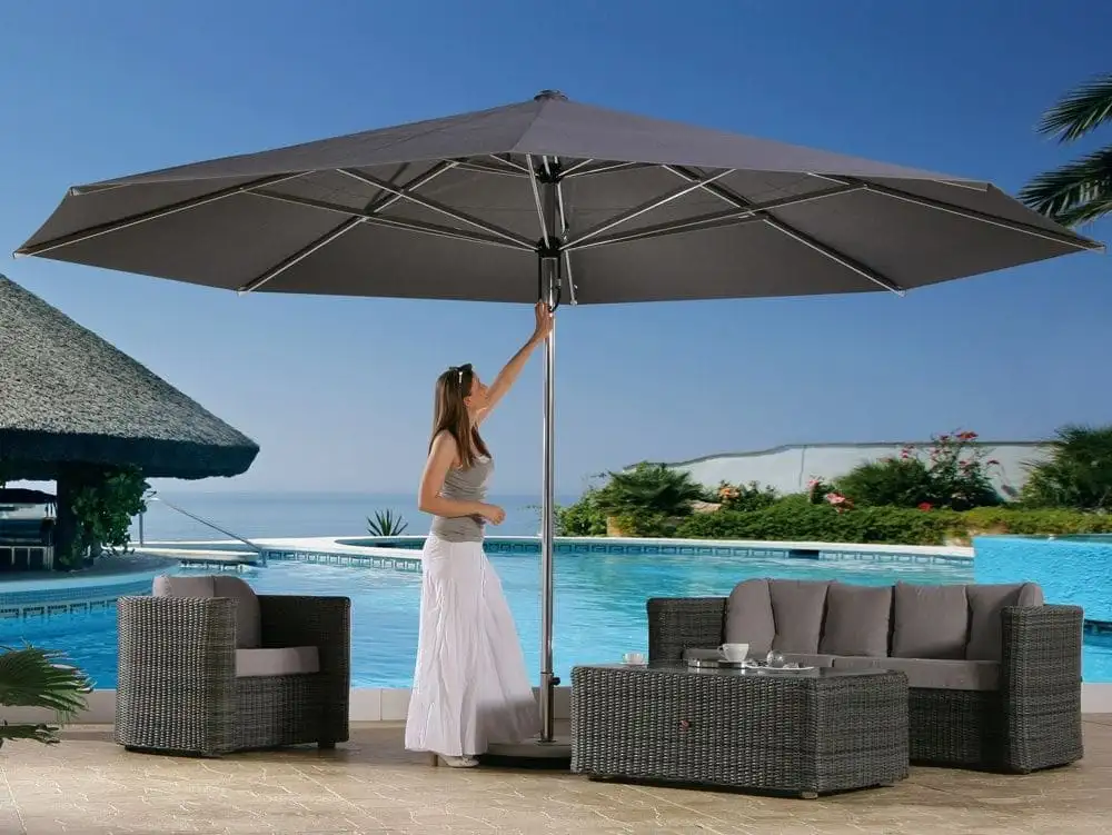 Une femme ouvre d’une main un grand parasol gris au bord d’une piscine, au-dessus d’un salon de jardin