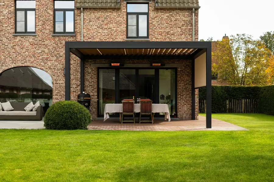 Pergola à lames anthracite adossée à une maison en briques, une table dressée dessous