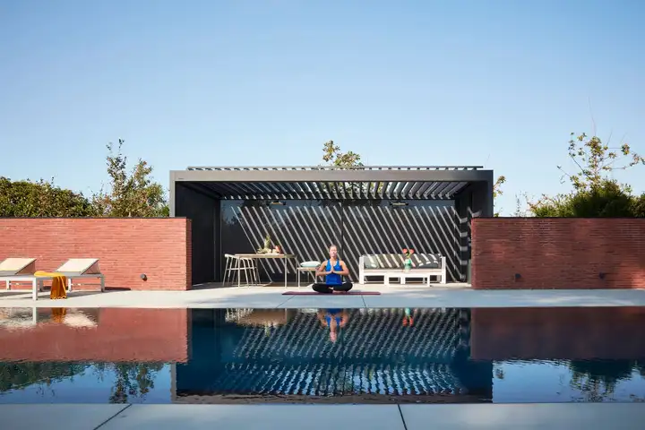 Pergola anthracite à lames au bord d’une piscine, une femme assise dessous, reflets dans l’eau