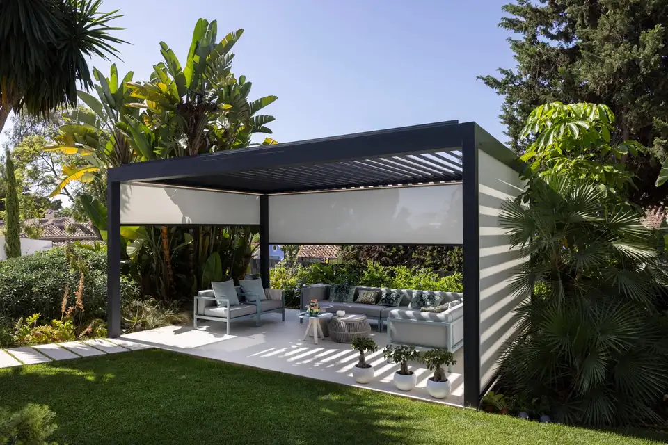 Pergola bioclimatique anthracite autoportée au jardin, au-dessus d’un salon d’extérieur, en partie fermée par des stores verticaux