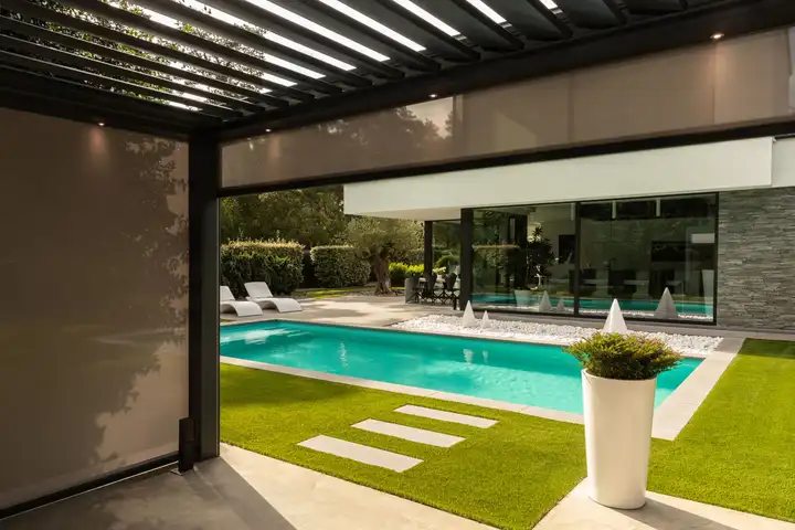 Pergola à lames fermée sur un côté par un screen, face à une piscine et une maison
