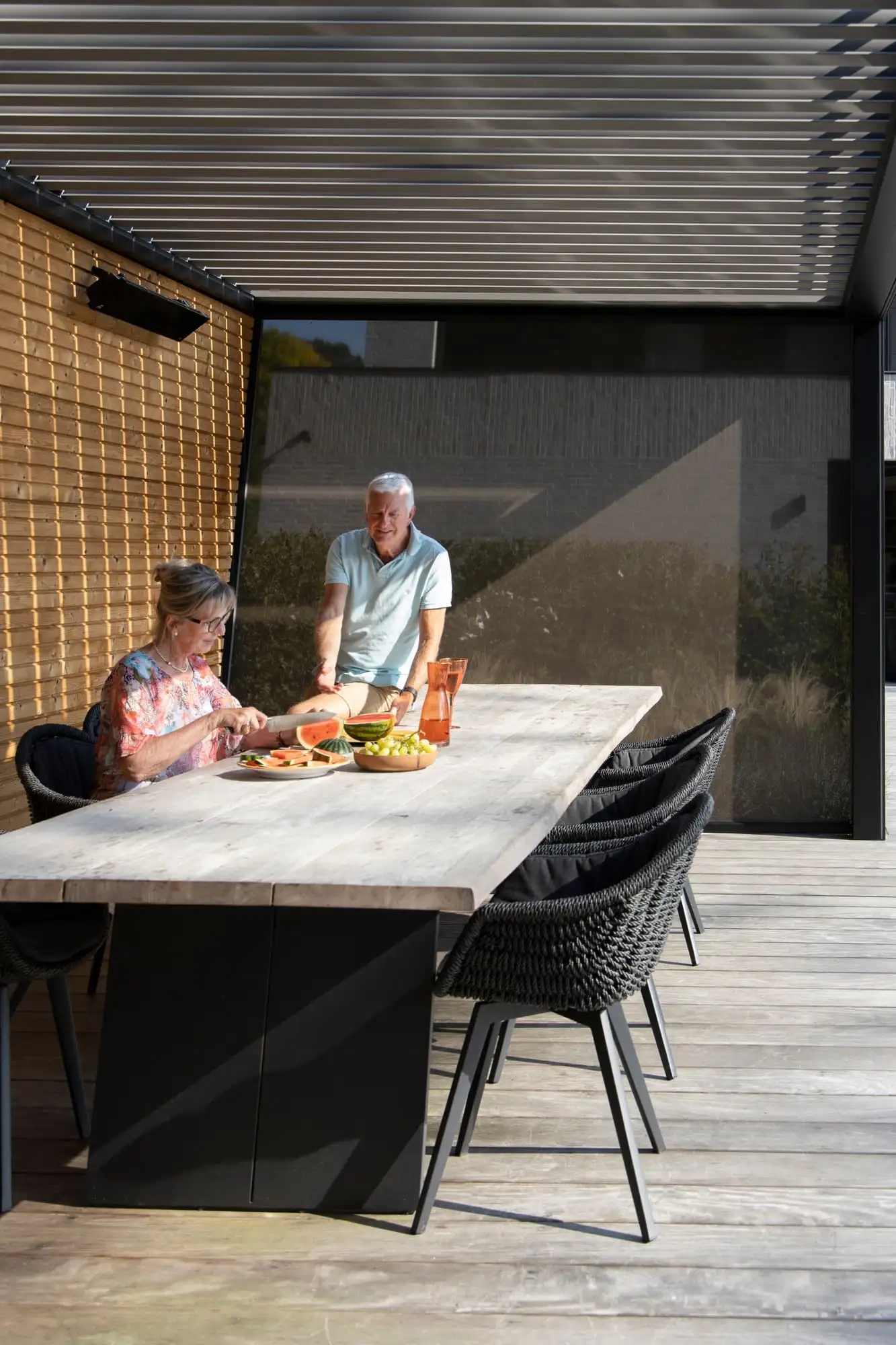 Un couple dresse la table sous une pergola bioclimatique anthracite, fermée sur un côté par un screen tressé