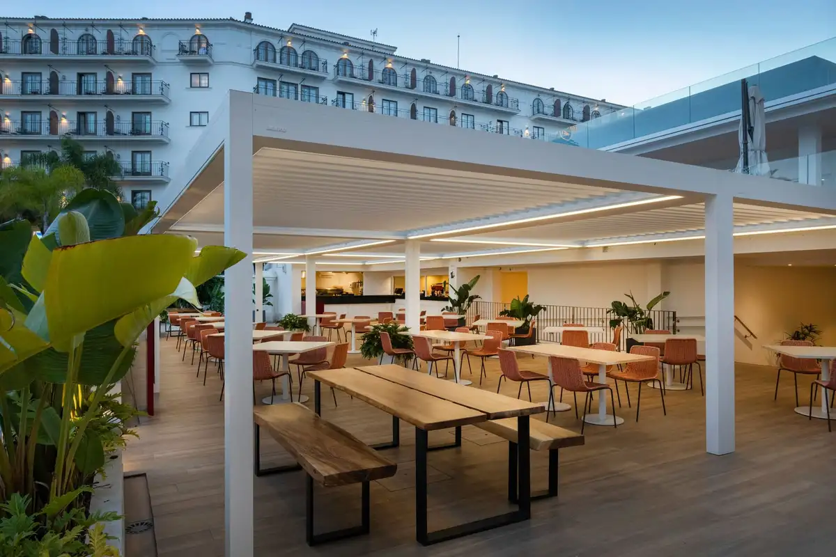 Grande pergola blanche à lames couvrant la terrasse d’un restaurant d’hôtel, tables et bancs en bois