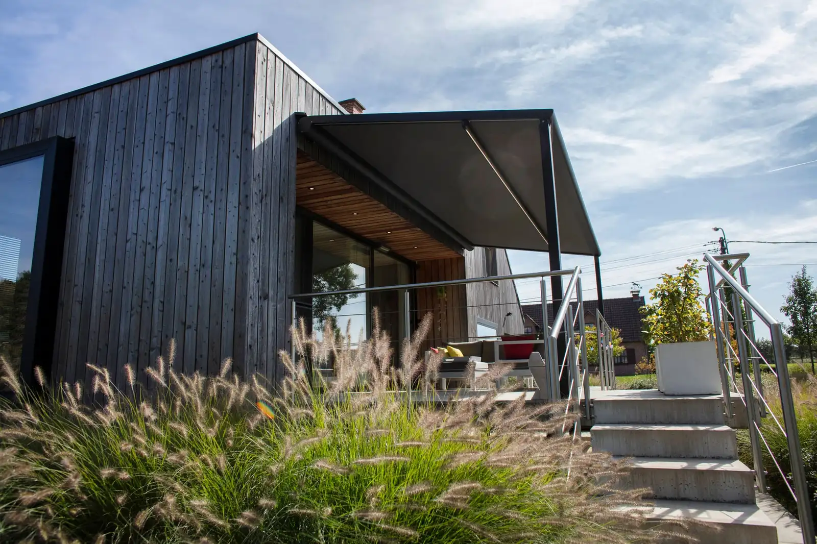 Pergola à toile anthracite adossée à une maison en bois sombre, au-dessus d’une terrasse surélevée