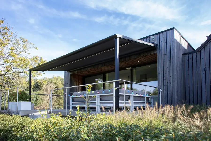 Pergola à toile anthracite adossée à une maison en bois, au-dessus d’une terrasse