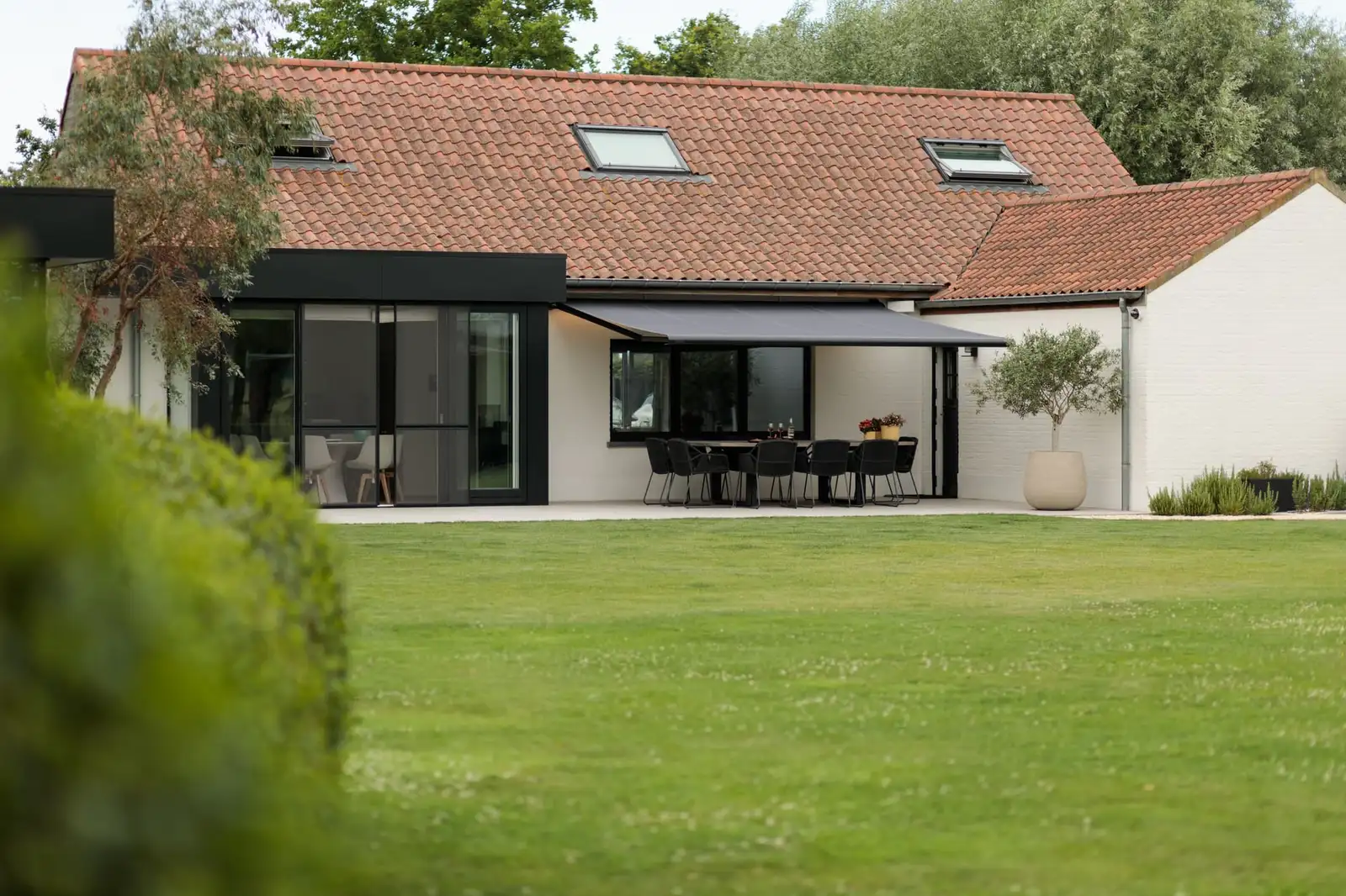 Store banne à toile gris anthracite déployé au-dessus de la terrasse d’une maison, entre ses deux ailes, face au jardin