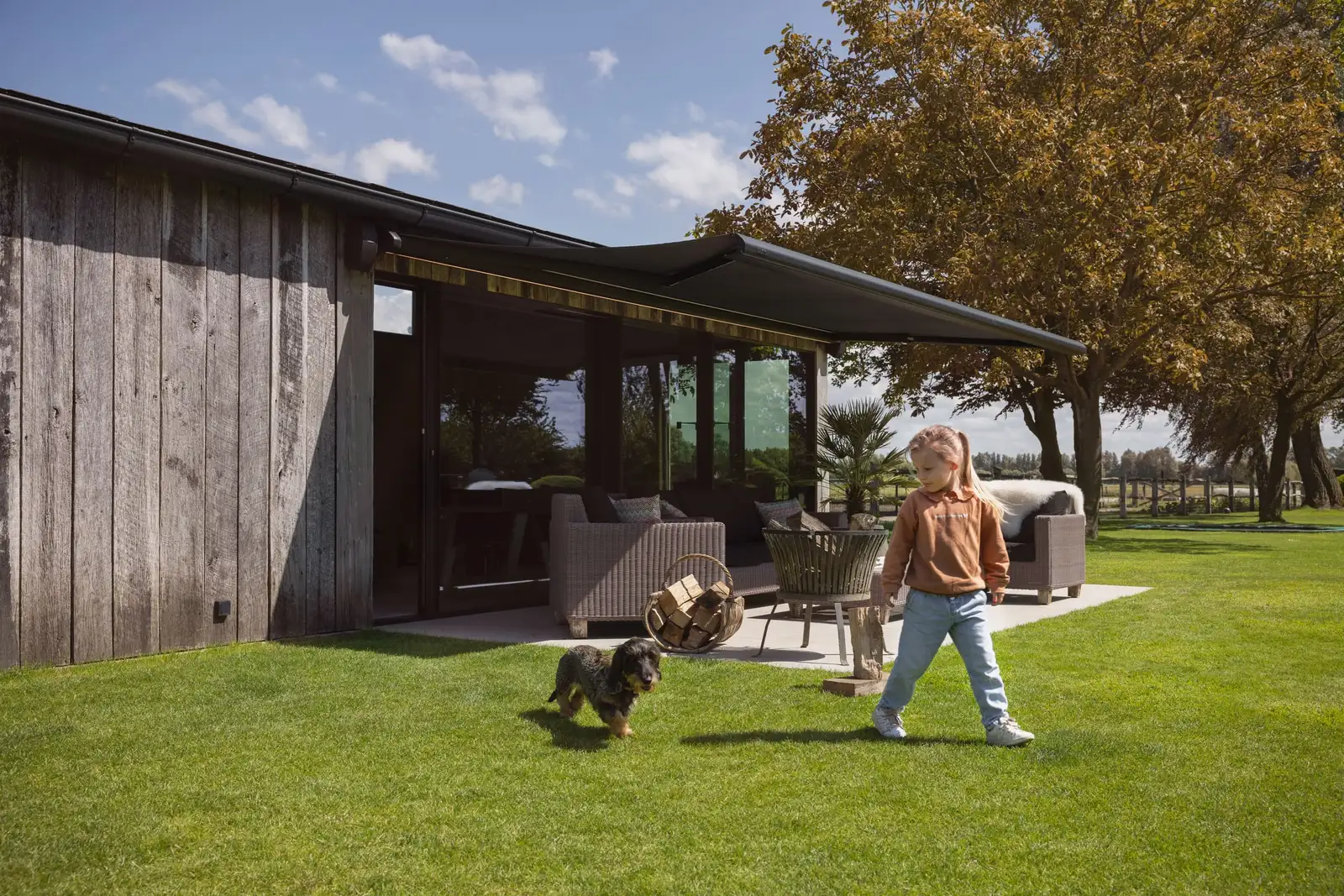 Une petite fille et son chien jouent sur la pelouse, devant la terrasse d’une maison abritée par un store banne anthracite