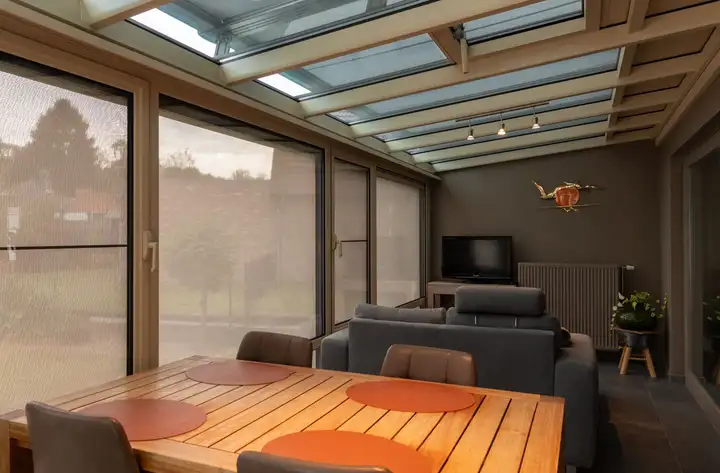 Intérieur d’une véranda en bois à la lumière tamisée par des stores, sur la toiture et devant les baies