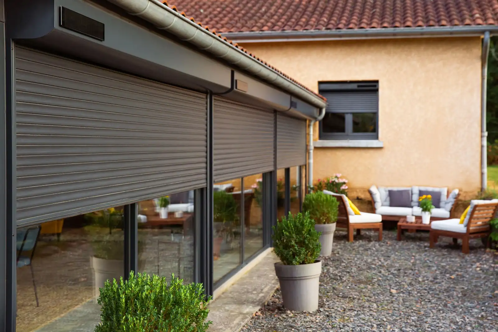 Volets roulants gris anthracite à demi baissés devant les grandes baies d’une maison, coffres posés sous l’avant-toit